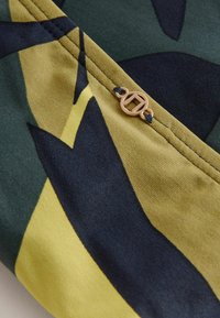 Grüner und gelber Stoff mit abstrakten Formen; verfügt über einen Akzent aus goldfarbenem Metall und Nähdetails. Weiche Textur mit matter Oberfläche.