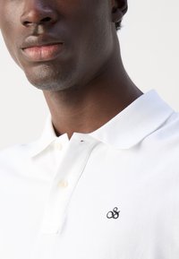 Hvit polo skjorte med knappet krage, med en subtil svart brodert logo på brystet og et strukturert stoff.