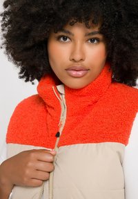 Giubbotto smanicato imbottito con parte superiore in fleece arancione e parte inferiore beige, dotato di zip frontale, colletto alto e una texture morbida.