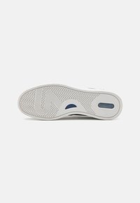 Weiße Sneaker-Sohle mit einem Fischgrätprofil, einer dicken Gummisohle und einem kleinen blauen Akzent nahe der Ferse.