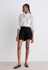 Camisa blanca transparente de botones con un estampado floral, combinada con pantalones cortos de tiro alto negros con lentejuelas y zapatos planos negros. Diseño simple y moderno.