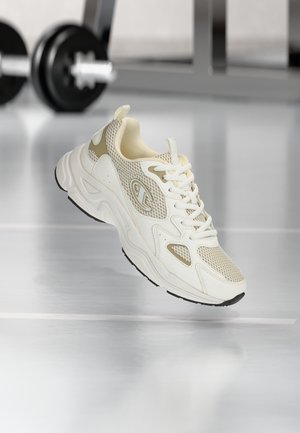 White and beige athletic sneaker with mesh and leather details, positioned mid-air on a reflective gym floor with blurred dumbbells in background.