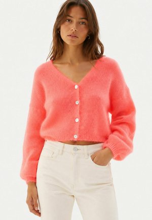 Jeune femme portant un cardigan boutonné duveteux corail vif avec des manches ballon et un pantalon beige taille haute, debout avec une main dans la poche.