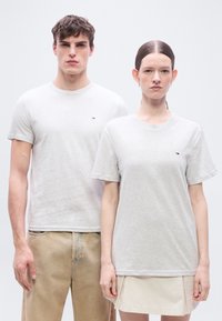 Δύο μοντέλα φορούν ανοιχτό γκρι T-shirts με μικρά λογότυπα στο στήθος, συνδυασμένα με μπεζ παντελόνια και μπεζ φούστα, στέκονται μπροστά σε ένα απλό φόντο.