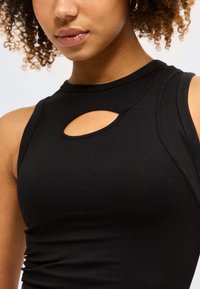 Γυναίκα με σγουρά μαλλιά που φοράει μαύρο αμάνικο τοπ με μικρή οβάλ κοπή κοντά στη λαιμόκοψη.