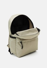 Mochila beige hecha de tejido texturizado, con cierre de cordón, solapa superior y forro interior negro. Se observa un bolsillo frontal.