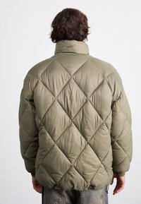 Padded olive green jacket with diamond quilted design, high collar, and elastic cuffs. Features "OBEY" embroidery on the neck.