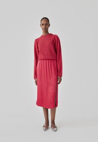 Rød strikket sweater med lange ærmer, parret med en lyserød silkesnor med sideslidser. Modellen bærer sølv høje hæle.