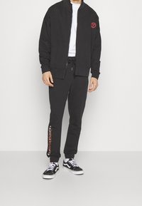 Schwarzer Reißverschluss-Sweatshirt mit strukturiertem Kragen, kombiniert mit passenden Jogginghosen mit einem roten Logo-Print am linken Bein. Getragen mit schwarzen Sneakern.