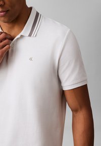White polo shirt with short sleeves, featuring a textured fabric, a two-button placket, and a gray-striped collar. Small logo on chest.