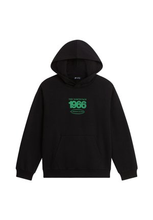 Dreng iført en sort hættetrøje, olivengrønne bukser, beige vandresko og en rød hue, der holder i en grøn rygsækrem og smiler.