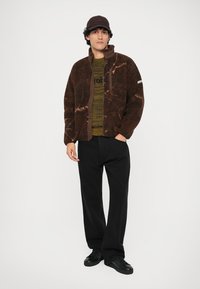 Καφέ ζακέτα από τεχνητή γούνα με ανοιχτόχρωμο μαρμάρινο σχέδιο, φορεμένη πάνω από ένα πράσινο πλεχτό πουλόβερ, συνδυασμένη με μαύρο παντελόνι και μαύρα παπούτσια.