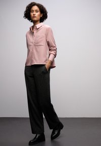 Blusa rosa chiaro con colletto e taschino sul petto, abbinata a pantaloni larghi grigio scuro e scarpe nere. Tessuto morbido e vestibilità rilassata.