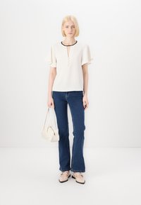 Bela majica kratkih rokavov z obročastim izrezom in tremi gumbi, v kombinaciji z modrimi flared jeans hlačami in majhno belo torbico.