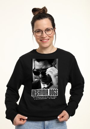 Femme souriante dans un sweat à capuche noir arborant un graphique de "Reservoir Dogs", tenant sa main contre des lunettes de soleil, se tenant devant un fond clair.