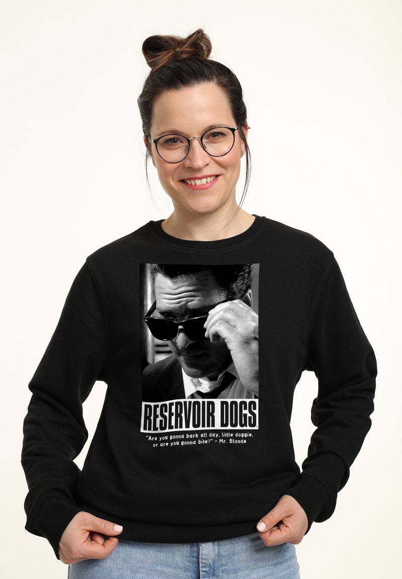 Lächelnde Frau in einem schwarzen Sweatshirt mit einem "Reservoir Dogs"-Grafikdesign, die ihre Hand an eine Sonnenbrille hält, vor einem hellen Hintergrund stehend.