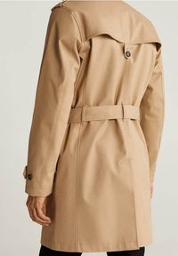 Beiger Trenchcoat mit tailliertem Design, großem Kragen und Knopfdetails. Er verfügt über eine glatte, wasserabweisende Textur und eine strukturierte Form.