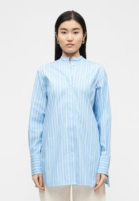 Γυναίκα με μακριά σκούρα μαλλιά που φοράει ανοιχτό μπλε και λευκό πουκάμισο με κάθετες ρίγες και λευκό παντελόνι, στέκεται μπροστά σε απλό φόντο.