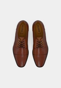 Chaussures habillées en cuir marron avec une finition polie, comportant six œillets, un bout pointu et des accents de couture décoratifs. Marque ALDO visible à l'intérieur.