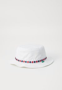 White bucket hat made of cotton. Features a decorative band with red, blue, and black diamond patterns. Soft texture, wide brim.