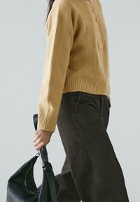 Maglione a coste color senape con patta con bottoni, abbinato a pantaloni marrone scuro. Una borsa in pelle nera testurizzata è tenuta in mano.