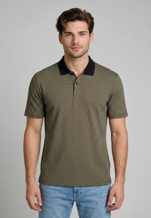 Homme aux cheveux courts et bruns portant un polo vert à manches courtes avec un col noir et un jean bleu clair, debout devant un fond gris.