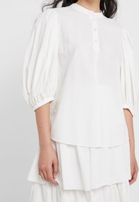 Blusa bianca, strutturata, con maniche a sbuffo, colletto alto e chiusura frontale con cinque bottoni. Il tessuto presenta righe verticali.