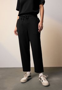 Juodi sportiniai kelnės su raišteliu juosmens srityje, pasižyminčios laisvu kirpimu ir lygiu paviršiumi. Derinami su smėlio spalvos sportbačiais ir baltais kojinėmis.