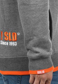 Szary sweter z pomarańczowymi akcentami, ściągaczami przy mankietach i bocznymi kieszeniami. Na przodzie haftowany tekst: "!SOLID" oraz "Since 1993".