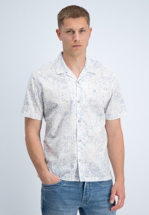 Jonge man met kort haar, gekleed in een wit overhemd met korte mouwen en een subtiel blauw patroon, en een blauwe spijkerbroek, staand tegen een effen achtergrond.
