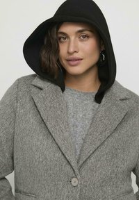Donna con capelli corti e mossi che indossa un cappuccio nero, un cappotto grigio testurizzato e un maglione a trecce grigio chiaro, rivolta verso di fronte con un'espressione neutra.