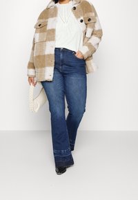 Brown and white checked fuzzy jacket with two front pockets, paired with a light top and dark blue flared jeans. White braided handbag.