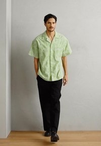 Short-sleeve, light green shirt with a sunburst pattern, paired with black trousers. Model stands on a wooden floor against a gray wall.