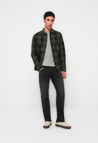 Ung mann som står mot en enkel hvit bakgrunn, iført en grønn og svart rutete jakke, grå skjorte, svarte jeans og hvite joggesko.