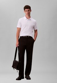 Tricou polo alb cu mâneci scurte, pantaloni negri, curea neagră și pantofi negri. Modelul ține o geantă de umăr neagră cu o curea.
