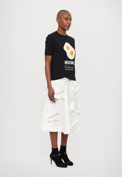 Zwarte t-shirt met ei-afbeelding en de tekst "Moschino", gecombineerd met een witte rok met gelaagde, gevouwen accenten. Zwarte hakken en sokken maken de outfit compleet.