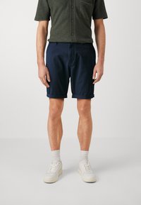 Pantaloni corti di cotone blu navy con risvolti, abbinati a una camicia verde scuro con chiusura a bottoni e taschino sul petto. Scarpe da ginnastica bianche indossate.