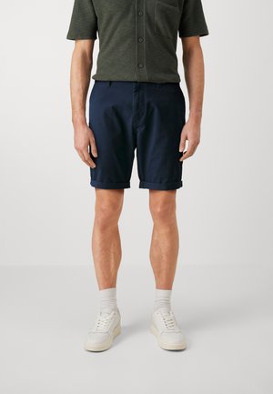 Pantalones cortos de algodón azul marino con puños doblados, combinados con una camisa verde oscura de diseño abotonado y bolsillo en el pecho. Se llevan zapatillas blancas.