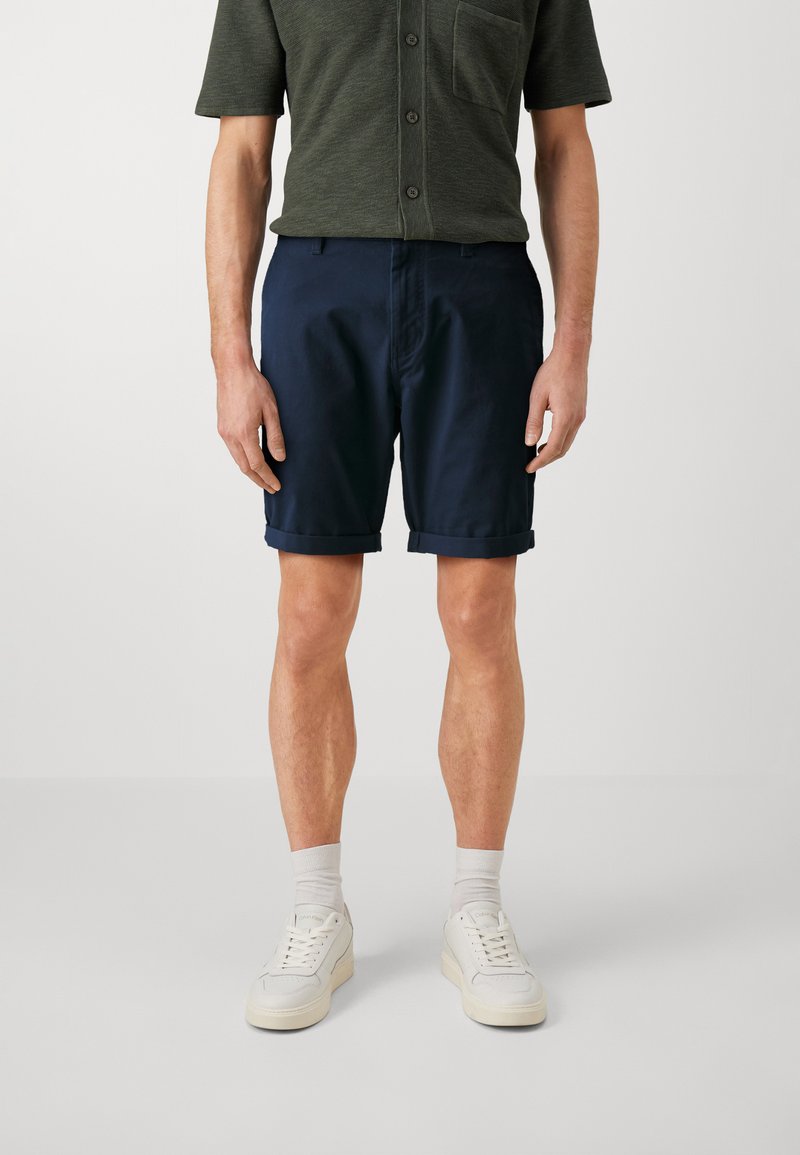 Pantaloni corti di cotone blu navy con risvolti, abbinati a una camicia verde scuro con chiusura a bottoni e taschino sul petto. Scarpe da ginnastica bianche indossate.