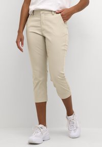 Beige cropped pants made of smooth fabric with a tailored fit, featuring side pockets and a cuffed hem, paired with white athletic sneakers.