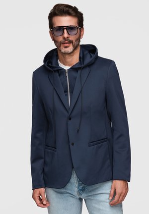 Homme portant un blazer bleu marine par-dessus un sweat à capuche zippé bleu marine, un jean bleu clair et des lunettes de soleil foncées sur un fond gris.