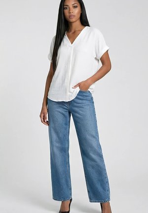 Kobieta z długimi włosami, ubrana w biały krótki top z dekoltem w serek i luźne niebieskie jeansy, stojąca z jedną ręką w kieszeni.