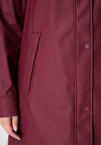 Manteau imperméable bordeaux au toucher lisse, avec une fermeture à pression, des poches latérales et des coutures discrètes.