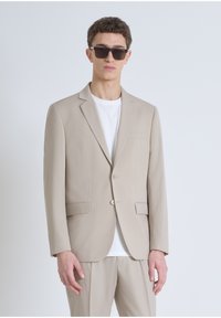 Veste de costume beige sur mesure en tissu, fermeture à un bouton, revers en pointe et poches avant. Portée sur un t-shirt blanc.
