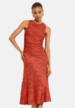 Kvinde iført en ærmeløs, tekstureret brændt orange midi-kjole med en tætsiddende talje og en udvidet nederdel, stående mod en ensfarvet baggrund.