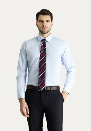 Camicia azzurro chiaro con maniche lunghe, colletto button-down, abbinata a una cravatta a righe rossa scura e blu navy, e pantaloni scuri con una cintura marrone.
