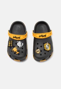 Svarta crocs med gula remmar, utrustade med dekorativa nålar: en grävling, en trofé och ett vapen. Texturerat material med ventilationshål och ett vadderat inre.
