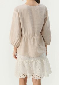 Vaaleanbeige pellavapusero, jossa on ilmavat hihat ja joustava vyötärö, yhdistettynä valkoiseen, pitsikoristeiseen hameeseen, jossa on kerrokselliset röyhelöt.