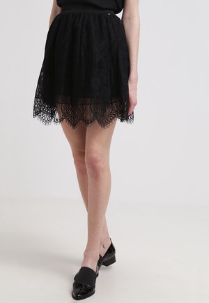 Kobieta ubrana w czarną koronkową spódnicę z falistym brzegiem i czarne lakierowane buty na niskim obcasie, stojąca na tle jednolitego tła.