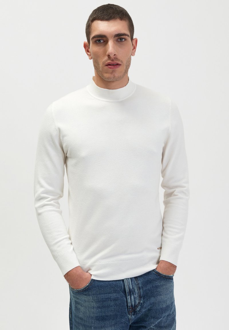 Maglione bianco a maniche lunghe con colletto alto, realizzato in morbido tessuto a maglia. Design aderente, con polsini e orlo a costine. Abbinato a jeans in denim blu.